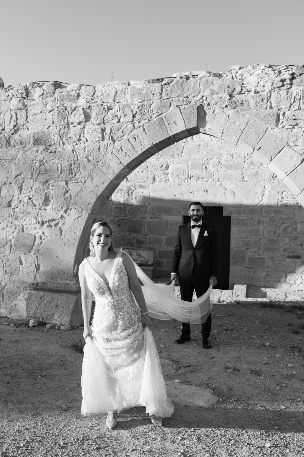 Black and white wedding portrait of Giorgos and Andriana beneath a stone arch